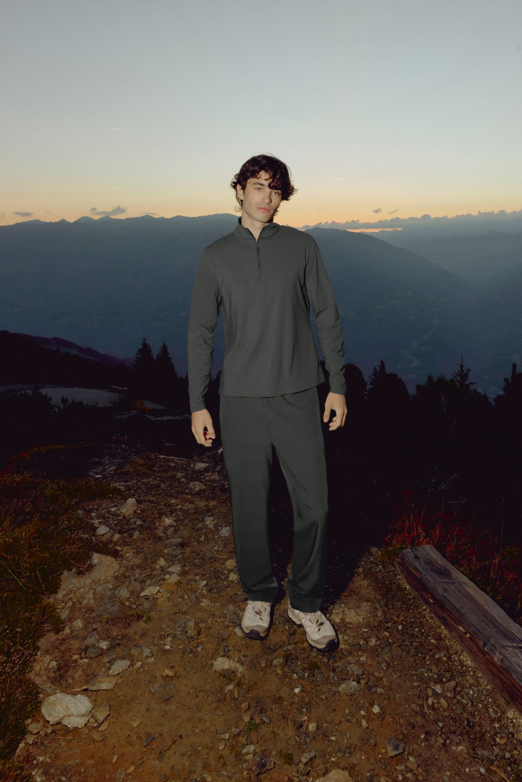 Man wearing olive green CASPARA Prime Core pants standing on a rocky hillside at dusk.